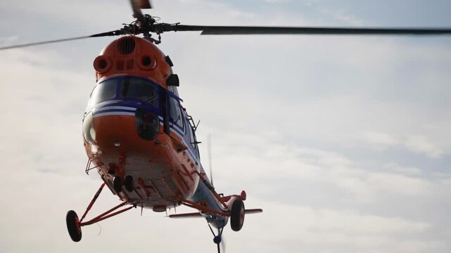 Low-altitude orange rescue helicopter hovers against a cloudy sky, rotating main rotor blades blur as a suspended cargo hook and line hang below during an aerial lift operation