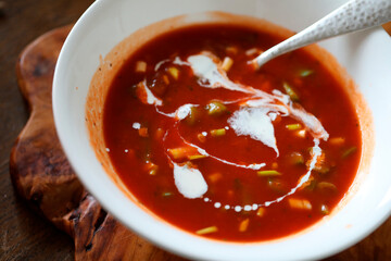 COLOURFUL TOMATO BASED VEGETABLE SOUP WITH STYLISH SWIRL OF PLANT BASED CREAM IN WHITE BOWL WITH BEATON METAL SPOON ON RUSTIC WOODEN BOARD .