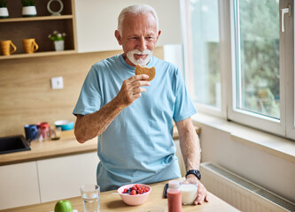 A senior man is eating a cokie energy bar and drinking milk in kitchen. Having a break after a morning exercise. The man is enjoying his meal, vitality, healthy eating and active senior concept