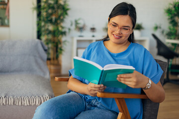 A young woman sits comfortably in a cozy chair, joyfully reading a book in a bright and inviting living room filled with greenery and artistic decor.