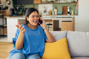 The excited young woman energetically chats on her phone, surrounded by a beautifully decorated living space, reflecting her vibrant personality and engagement.