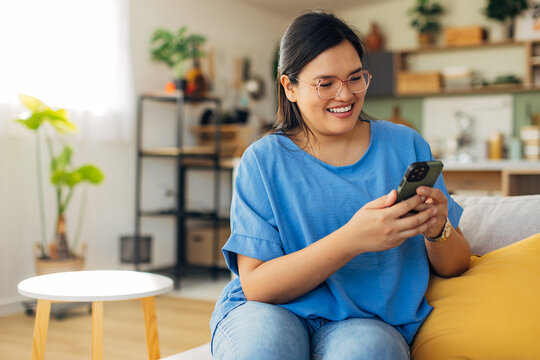 A young woman wearing casual clothing and glasses smiles brightly into her phone, displaying energetic expressions in a lively and well-decorated living room setting.