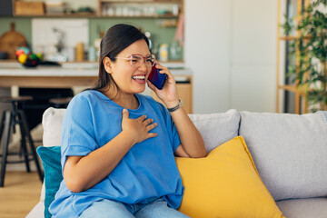 A woman laughs joyfully while talking on the phone, comfortably seated on a couch adorned with a yellow pillow, creating an inviting and cheerful living space ambiance.