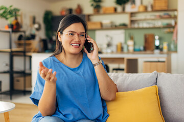 A joyful young woman is seen smiling and gesturing while talking on her phone in a cozy living room, reflecting a modern and vibrant lifestyle filled with positivity.