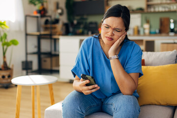 A young woman appears stressed while looking at her smartphone, portraying the impact of digital communication on personal emotions in a cozy home environment.
