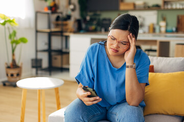 A young woman appears stressed while using a smartphone in her cozy living room, illustrating the emotional impact of technology on mental well-being and relaxation.
