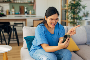 A young woman joyfully engaging with someone through her smartphone during a video call while seated comfortably in a cozy living room space full of greenery.