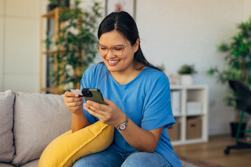 A cheerful woman utilizes her phone and credit card for online shopping, capturing the essence of modern retail experiences in a cozy and stylish living room ambiance.