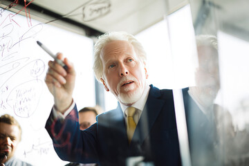 Senior businessman explaining strategy confidently on glass board in modern office, sharing ideas with colleagues during meeting, focused and professional atmosphere
