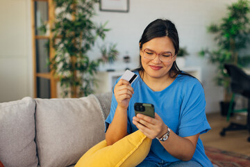 An enthusiastic young woman is holding her credit card and phone, smiling as she engages in online shopping, enjoying the simplicity of making purchases at home.