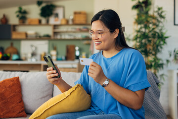 In a chic home decor, a smiling woman engages in online shopping, holding her credit card and smartphone, symbolizing convenience and joy in the digital age.
