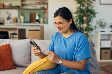 A delighted woman sits comfortably in her stylish living room, using her smartphone and credit card to shop online, showcasing a perfect blend of comfort and technology.