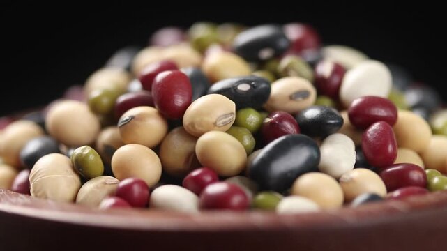 Pile of five mixed beans (black, red, green, white, and soybeans in a brown wooden bowl rotating on black background. Close-up shot