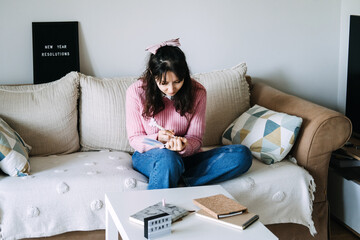 Young adult woman sits on sofa writing goals in small notebook next to letter board displaying NEW...
