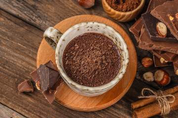 Hot chocolate topped with grated dark chocolate in a ceramic mug on a rustic wooden background, surrounded by chocolate pieces, nuts and cinnamon, creating a warm cozy dessert atmosphere