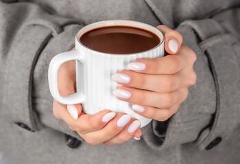 Woman holding a warm cup of hot chocolate in both hands, close-up view of white ceramic mug and manicured fingers in a cozy grey coat, conveying warmth, comfort, winter mood