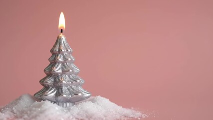 A silver Christmas tree candle with a lit flame, standing on a bed of artificial snow against a soft pink background, creating a festive and cozy holiday atmosphere. - Powered by Adobe