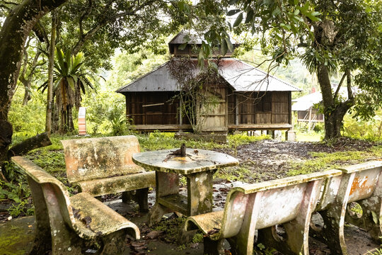 Old moss-covered stone benches and table beneath lush trees facing the historic Masjid Lama Papan, a traditional wooden mosque in Papan Heritage Village, Perak, Malaysia