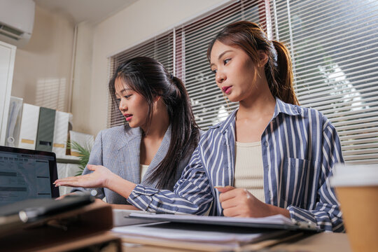 Two young women collaborate, discussing business project details on laptop screen, working from home office, team learning online strategy in modern remote environment