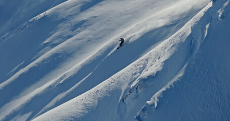 Aerial photo of a free ride skier carving through fresh powder snow on a steep alpine slope. Drone shot capturing the thrill of first tracks in pristine winter mountains.