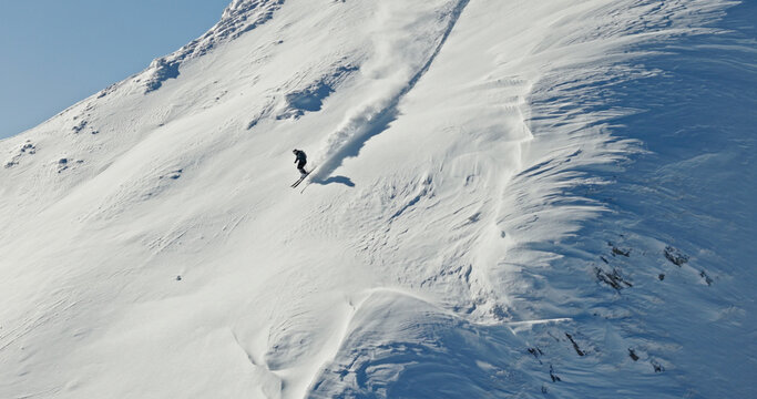 Aerial photo of a free ride skier carving through fresh powder snow on a steep alpine slope. Drone shot capturing the thrill of first tracks in pristine winter mountains.