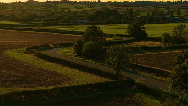 Blue car curving along countryside road in golden sunset fields. Road driving, car traveling, countryside lifestyle vacation. Compact vehicle navigating rural winding roadway during evening glow