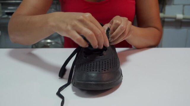 A woman in a red top laces up a black sneaker. She carefully threads the laces through the eyelets while sitting at a white table indoors.
