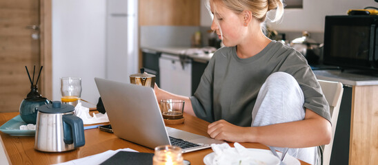Young woman working remotely at home kitchen table with laptop, coffee, and breakfast, representing...