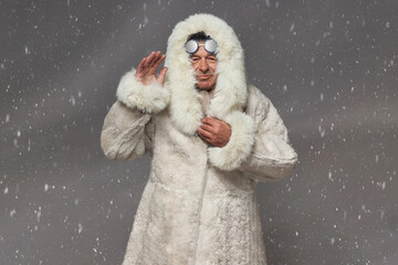A man in a luxurious white fur coat with a hood and glasses for protection from snow, dynamically waves his hand against the background of snowfall.