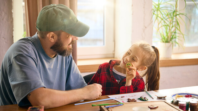 Daughter proudly holding painted clay smiley face while father watches. Concept of emotional encouragement, positive learning outcomes, playful art practice, and confidence-building crafting. - Powered by Adobe