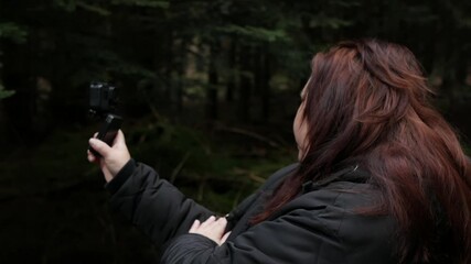 A side-profile shot of a female blogger or hiker in a dark jacket holding an action camera up to film herself against a dark, dense pine forest background. 