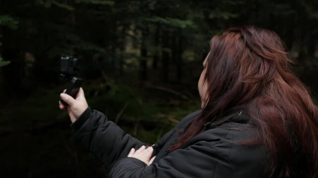 A side-profile shot of a female blogger or hiker in a dark jacket holding an action camera up to film herself against a dark, dense pine forest background. 