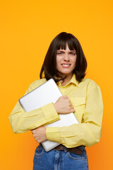 Fototapeta premium A young woman in a yellow shirt hugs a laptop against a bright orange backdrop, presenting a casual, energetic vibe perfect for tech, education, or creativity themes.