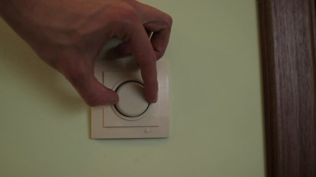 Male hand turning a round knob on a modern wall dimmer switch. Adjusting the brightness of the light and saving energy at home or office