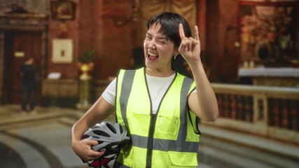 Young woman with short hair holding a bicycle helmet makes rock horns gesture in church interior while wearing a yellow high visibility vest; playful defiance.