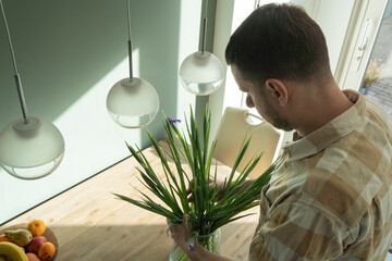 A man places a freshly arranged vase of irises on his kitchen table, completing the scene.  A centerpiece that enhances the warm, inviting atmosphere of his modern and thoughtfully decorated home