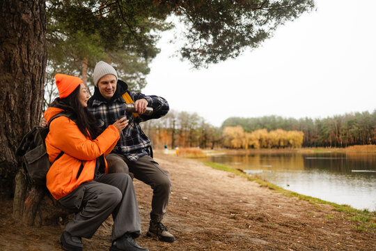 Hiking couple enjoys nature taking a break with water bottle or hot drink tea near lake in a lush forest during a cold day.