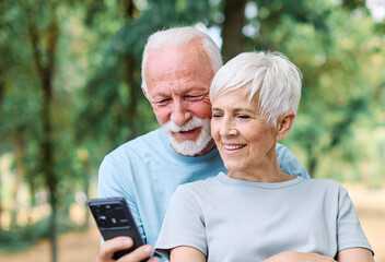 Happy active senior couple having fun using smartphone and wearing sportswear, after having an exercise sport activity outdoors