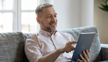 Smiling man on couch using tablet, relaxed at home with natural light and casual shirt, comfortable lifestyle and digital leisure mood