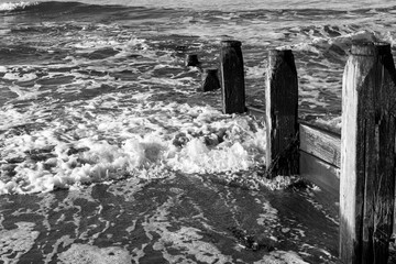 Waves breaking around weathered wooden groynes in black and white