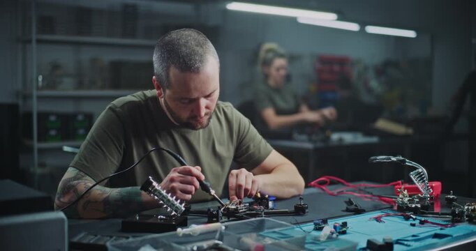 Adult Male Worker Closely Soldering Small Drone Component on Workbench, With Colleagues Working in Blurred Background. Concept of Collaborative Environment of Technology Production Facility.