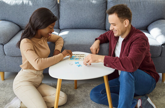 Young cheerful couple playing tic tac toe game together sitting on floor at home. Smiling man and african american woman playing board game in living room. Hobbies and leisure concept.