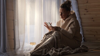 Woman wrapped in blanket sits by window with glass of wine scrolling through her phone Calm light blends silence with glow of screen Phone combines calm symbol of mindful telephonic contemplation