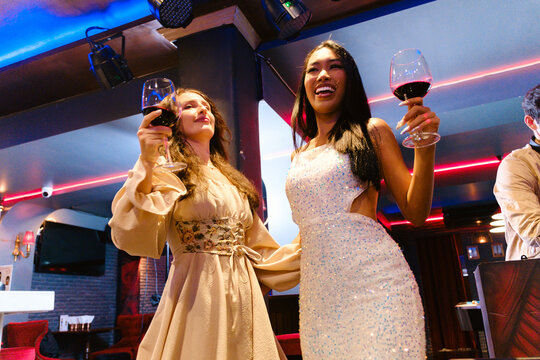 Two women in elegant attire enjoy wine and celebrate in a vibrant nightclub or lounge, smiling under party lights and sharing a festive, glamorous moment of friendship and joy.