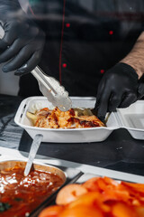 Close-up of shawarma ingredients in a to-go container, showing sliced meat, fresh vegetables, and white sauce being added, highlighting vibrant colors, textures, and appetizing details.
