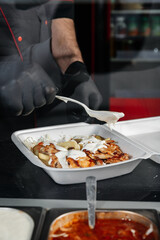 Close-up of shawarma ingredients in a to-go container, showing sliced meat, fresh vegetables, and white sauce being added, highlighting vibrant colors, textures, and appetizing details.
