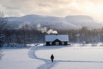 Winter landscape with a cozy cabin and a person walking towards it in snowy mountains