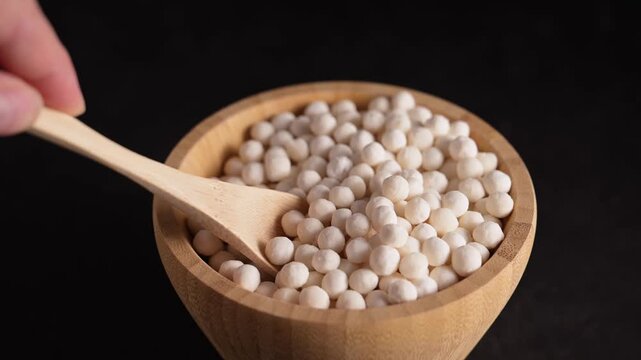 Hand scooping dry tapioca pearls with wooden spoon from bamboo wooden bowl. Raw tapioca starch balls made from cassava root, used for bubble tea and desserts.