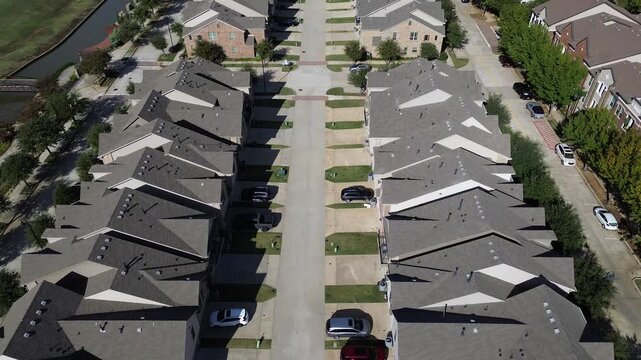 Symmetrical rows of new-build houses with dark shingle roofs, compact yards line central street near Riverside Drive, Flower Mound. Scenic canal, pedestrian bridge walkability, visual rhythm, TX