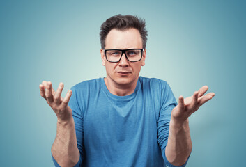 Naklejka premium Confused man in glasses raising his hands in a questioning gesture, expressing frustration or disbelief. Casual blue t-shirt, puzzled facial expression. On blue background.